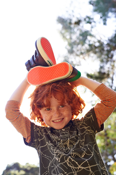 smiling boy with red curly hair holding shoes on his head with sunbeam coming trough them