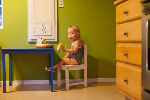 A blonde toddler girl in bathing suit holds a banana in one hand as she sits in a small chair at a small blue table with the other.