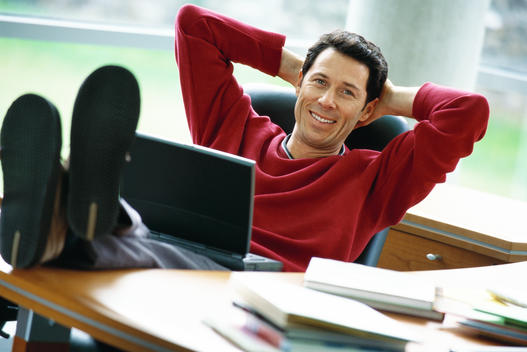 Man sitting at desk with feet up, holding laptop on lap, hands behind head