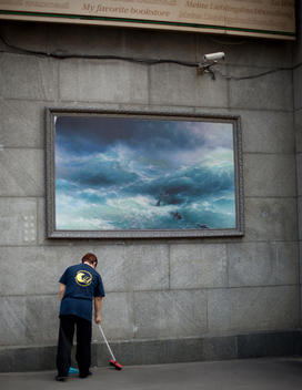 A janitor sweeping the street under a surveilance camera and a painting of a wreck at sea, Moscow, Russia.