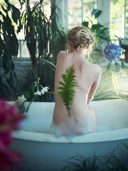 Young woman sitting on edge of bathtub, leaf stuck to her back, rear view
