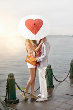 Romantic young couple behind heart umbrella with arms around each other on lakeside