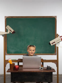 A girl sits at a school desk with a laptop and has cameras pointing at her from both sides