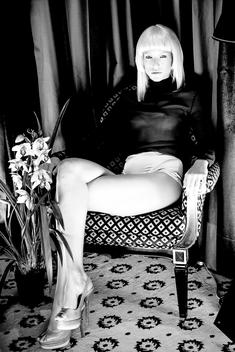 Sexy blond girl, sitting in opulent room with flowers , with see-through top and high heels.