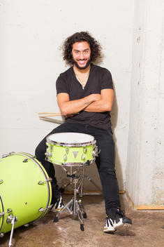 Mixed ethnicity millennial man in loft sitting smiling sitting in front of green drum set.