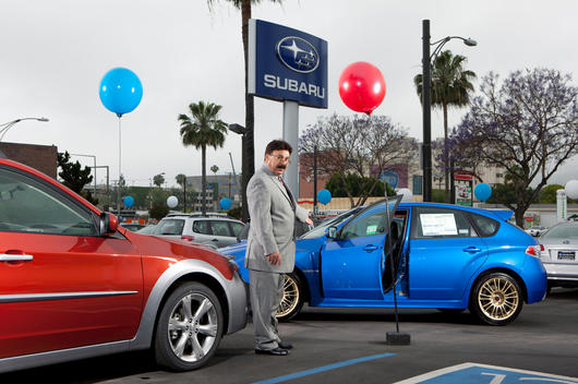 In the lot of a car dealership, a salesman stands between many new cars.