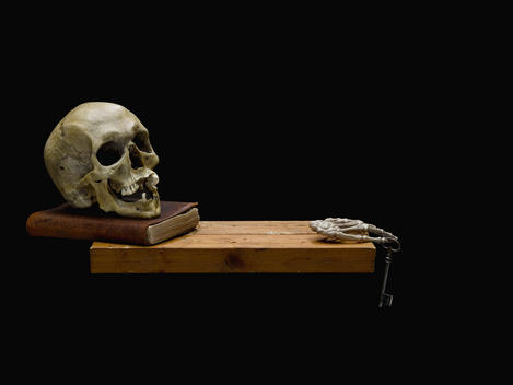 Still life with skull lying on a book and skeleton of a hand with key on black background.