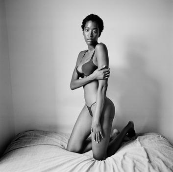 Dramatic black and white portrait of Fanta, a shy, beautiful African fashion model with a slim waist kneeling on a bed in bra and thong panties in window light in empty room. Brooklyn, New York