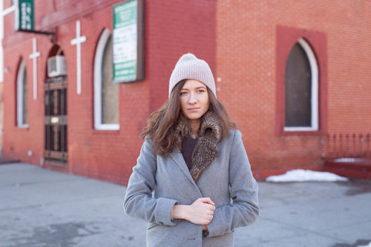 A pretty red-head Russian woman and model in a beanie/cap clenches her fur jacket in front of a colorful African-American/black church on a cold winter day during a street fashion shoot in Bed-Stuy, Brooklyn
