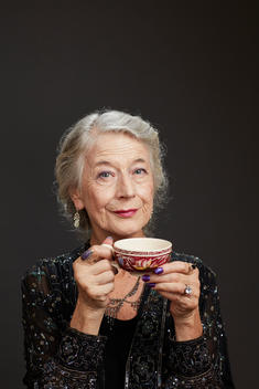 A formally dressed old woman holds a teacup with both hands to her mouth