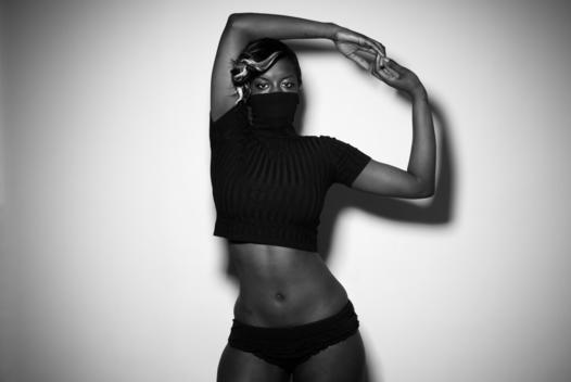 Young Black African-American Woman Wearing A Black Top That Covers Her Mouth And Nose And Panties Poses With Arms Above Her Head Against A White Backdrop. New York, Ny