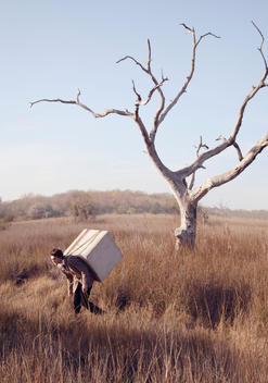 A man wandering through a marshland with a large concrete block strapped to his back while passing a dead tree