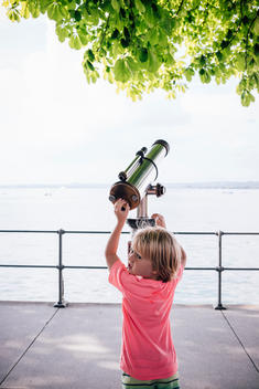 3 year old blonde boy in pink shirt playing with public telescope by Lake Constance in Austria.