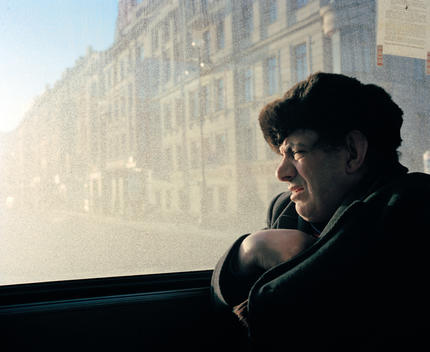 An Old Man Wearing A Coat And Fur Hat Sits On A Bus Looking Out Of The Window As The Sun Streams Through.