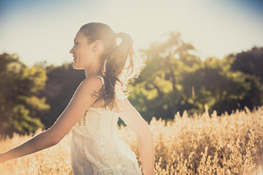 young woman running at summer through a cornfield wearing a light dress and smiling, back light and lens flare