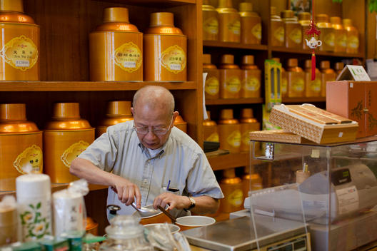 An Old Chinese Man Measures Tea Behind The Counter Of His Store.