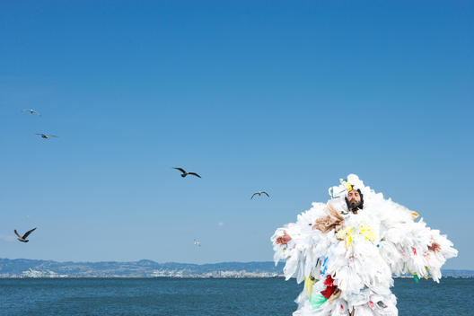 A man covered in plastic bags meditates in front of the ocean as seagulls fly around