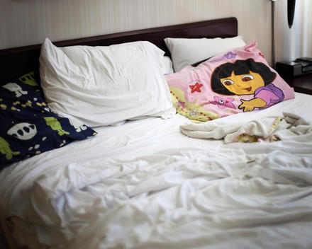 A hotel bed with white sheets and a blue, white and green alien spacemen pillow and a purple and pink Dora the Explorer pillow.