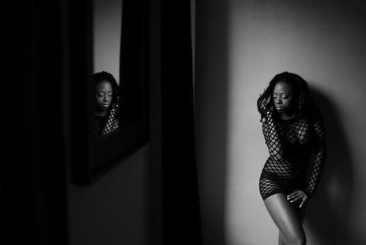 Young black African-American woman wearing black mesh fishnet body suit and lingerie whose face is reflected in a mirror stands against the wall in a dark room. Washington Heights, NYC