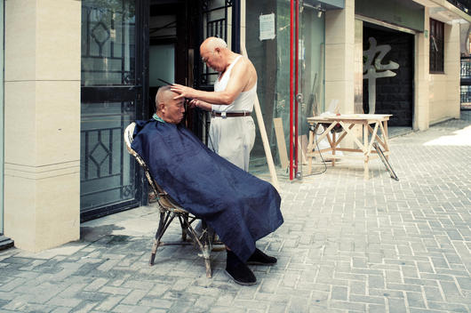 An old Chinese barber cutting hair for an old Chinese man outside the house.