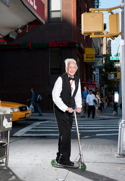 A very old waiter rides his scooter down a city sidewalk