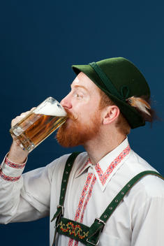 A bearded man wearing lederhosen drinks beer from a mug