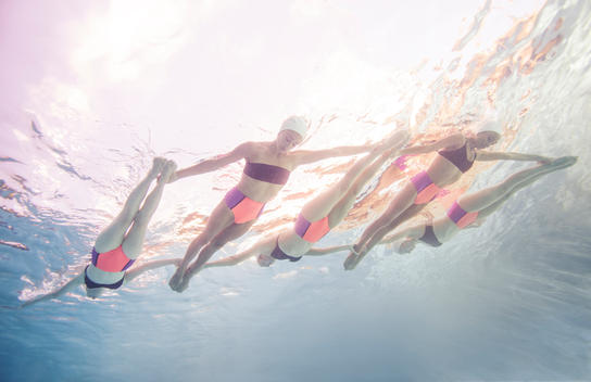 Five synchronized swimmers underwater on the surface facing down with legs straight and arms out, in a diagonal pattern