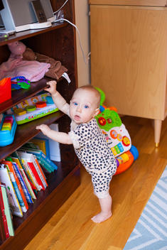 A baby stares up at the camera, leaning against a shelf full of colorful toys.