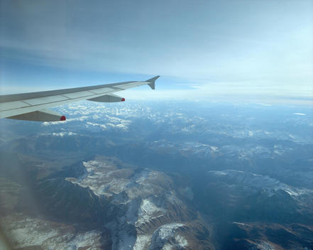 Flying Over The Alps, Mountains Are Only Partially Covered With Snow, The Wing Of The Airplane Leads On The Left Side Into The Image.