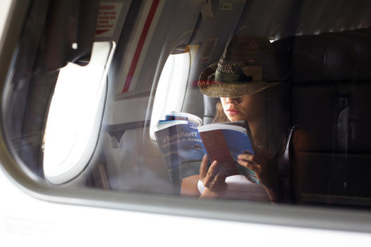 The Reflection Of A Young Women Wearing A Hat Reading A Book On A Plane