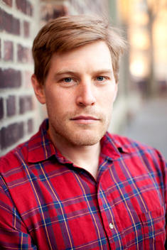 A portrait of a man looking forward with a stern expression wearing a red and blue plaid shirt.