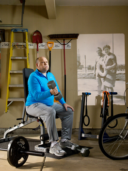 A man sits on an exercise machine holding a weight in his garage with an old family photo projected on the wall behind him