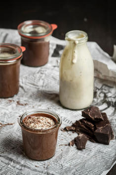 Jars of chocolate pudding with vanilla sauce in bottle on newspaper, close up