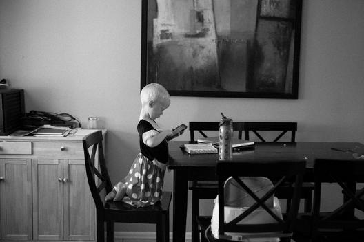 A young boy wears his big sister\'s polk-a-dot princess dress as he plays on his mother\'s cell phone at the kitchen table with a Mac computer and Book of Mormon scriptures nearby. Denver, Colorado