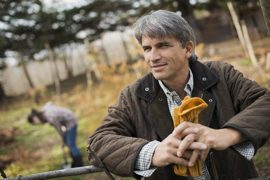 A man in a brown jacket, holding leather work gloves on an organic farm. A person digging in the ground with a spade.