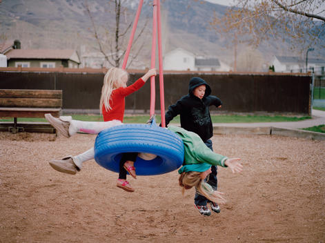A group of kids who are family, brother and sisters, play on a tire swing at Discovery Park on a cold spring afternoon near their suburban home with Wasatch Mountains in background. Pleasant Grove, Utah