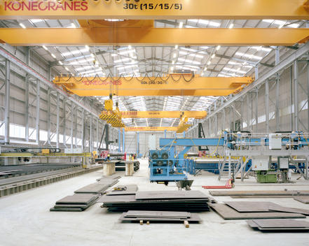 Large grey factory hall with a series of yellow cranes in the roof and steel sheets piled in the foreground.