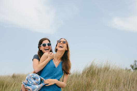 Two young female friends carrying picnic blanket laughing on dunes