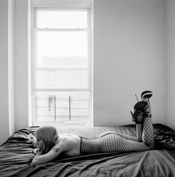 Young blonde busty large breasted semi-nude Russian woman with tattoos wearing a fishnet body suit and high heels sits on a bed alone. Bed-Stuy, Brooklyn, New York