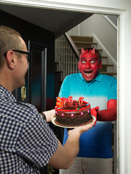 A normal guy hands a cake to a man dressed up as a devil at his front door