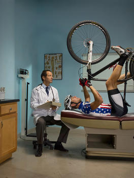 A bicyclist lies on his back on a table in an exam room holding his bike in the air while a doctor sits beside him and writes notes