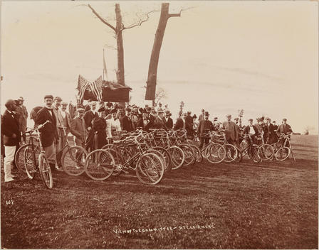 A Group Portrait Of Many People Standing Next To Bicycles Outdoors A Tent And Flags In The Background.