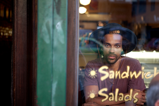 At an outdoor cafe shot through a storefront window using natural light, a portrait of hipster athletic black male model in urban clothing looking into camera