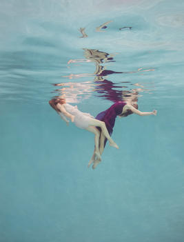 Two woman underwater in pink and maroon dresses with faces out of the water, floating away from each other