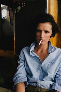 Serious portrait of a brown haired girl smoking a cigarette in a denim shirt.