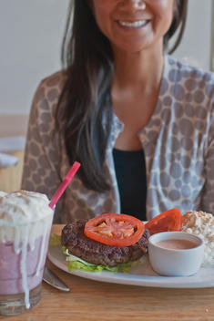 Smiling Woman Sitting At A Table With Plate Of High Protein Burger, Cottage Cheese Meal And A Milkshake.