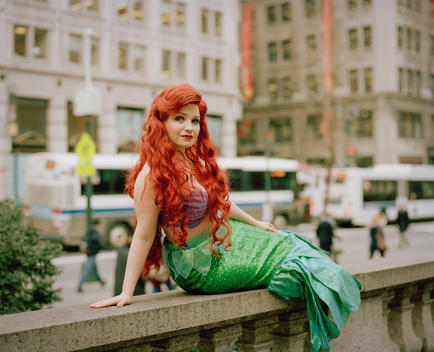 Portrait of Brittney, an ordinary woman who works as and impersonates Disney Princess Ariel from the Little Mermaid sitting on a marble ledge in front of the Public Library with traffic in background. New York, NY