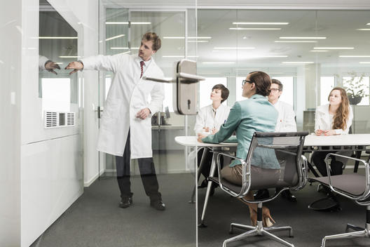 Man wearing lab coat pointing to screen in conference room, four colleagues watching