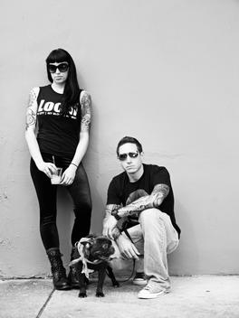 Woman and man posing against wall with a black dog.