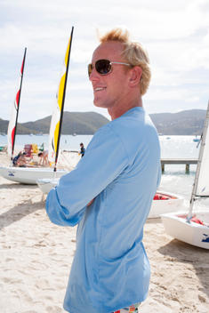 man with sunglasses stands with arms crossed on beach with sailboats in background
